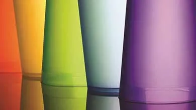 Close-up of colorful, translucent tumblers in orange, yellow, green, light blue, and purple, arranged in a row on a reflective surface.